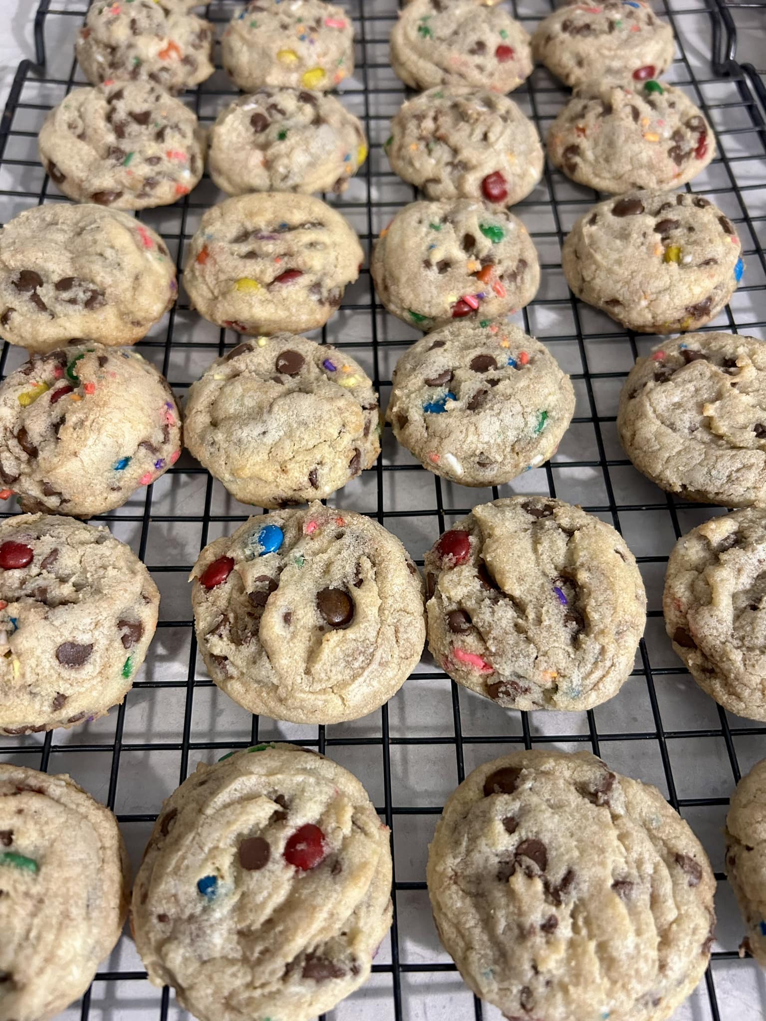 A large batch of freshly baked Soft Mini M&M Cookies cooling on a black rack.